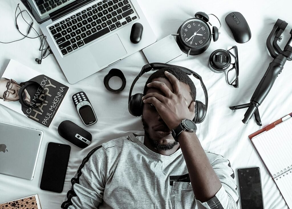 A young man lying on a bed surrounded by modern gadgets, books, and tech devices, illustrating digital lifestyle.