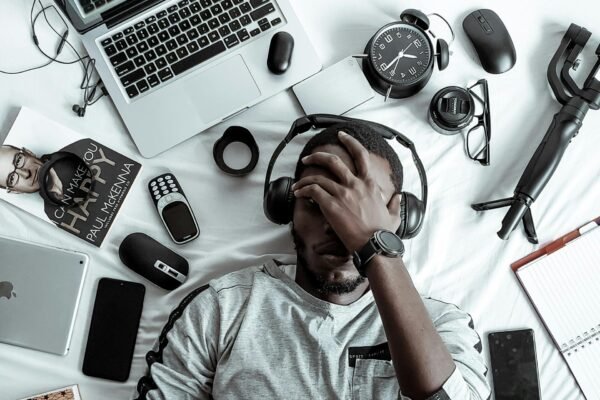 A young man lying on a bed surrounded by modern gadgets, books, and tech devices, illustrating digital lifestyle.